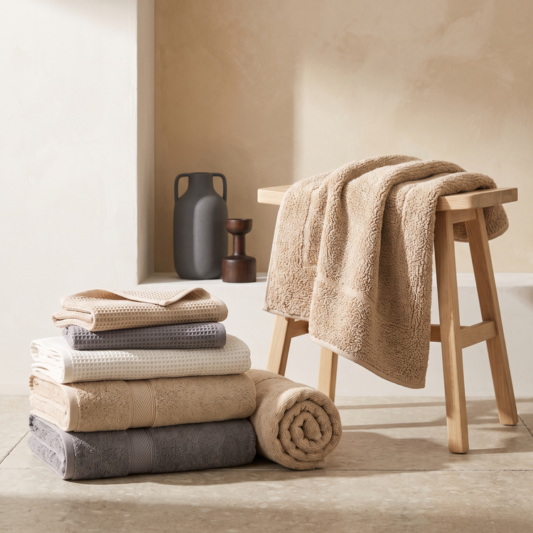 Folded waffle and terry towels in neutral tones beside a plush bath mat on an oak stool
